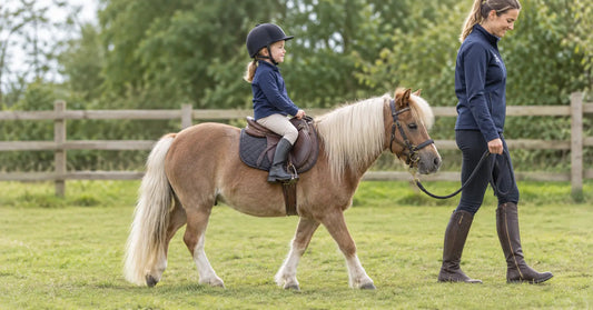 Child in a riding hat sitting in a lightweight treeless saddle on a miniature horse, led by an adult